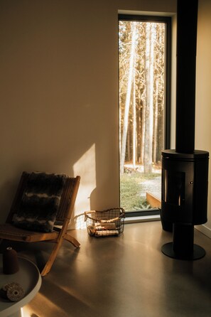 Ein gemütliches Zimmer mit einem großen Fenster, einem Korbstuhl, einem kleinen Tisch und einem Kaminofen.