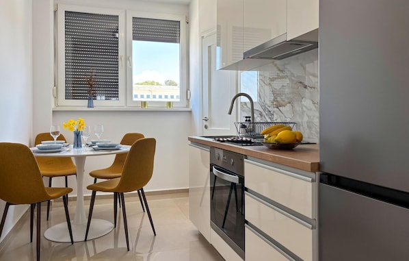 A modern kitchen with a dining area, a window with blinds, a stainless steel sink, and a refrigerator.