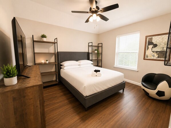 A bedroom with a bed, a ceiling fan, a TV, a bookshelf, a window with blinds, and a chair.
