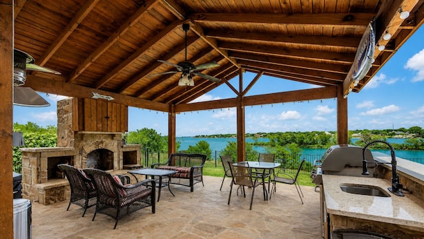 An outdoor patio with stone flooring, a stone fireplace, wicker furniture, a grill, and a view of a body of water. - Lakefront Oasis w/covered Patio + outdoor Kitchen (Fort Worth)