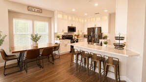 A modern kitchen with white cabinetry, a central island with bar stools, a dining table with chairs, and a 'let's eat' sign above the windows. - Lakefront Oasis w/covered Patio + outdoor Kitchen (Fort Worth)