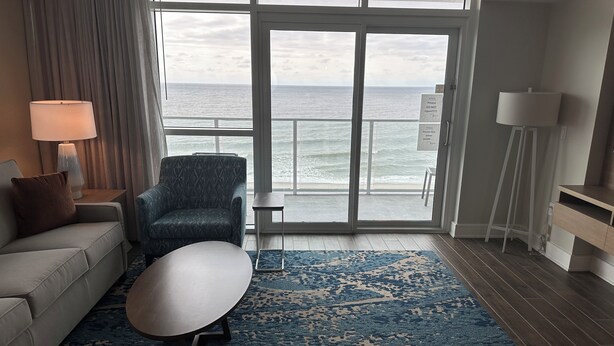 Habitación de hotel con sofá, sillón, reposapiés y vista al océano a través de una puerta corrediza.