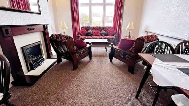 Ein Zimmer mit Kamin, einer Couch, zwei Sesseln, einem Esstisch mit Stühlen und einem großen Fenster mit Vorhängen.