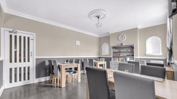 A dining area with a long table, multiple chairs, a chandelier, and a kitchen area with shelves. - Mallow View Guest House | Cleethorpes Beachside (Cleethorpes-Mallow View Guest House)