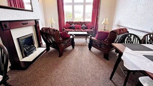 Ein Zimmer mit Kamin, einer Couch, zwei Sesseln, einem Esstisch mit Stühlen und einem großen Fenster mit Vorhängen.