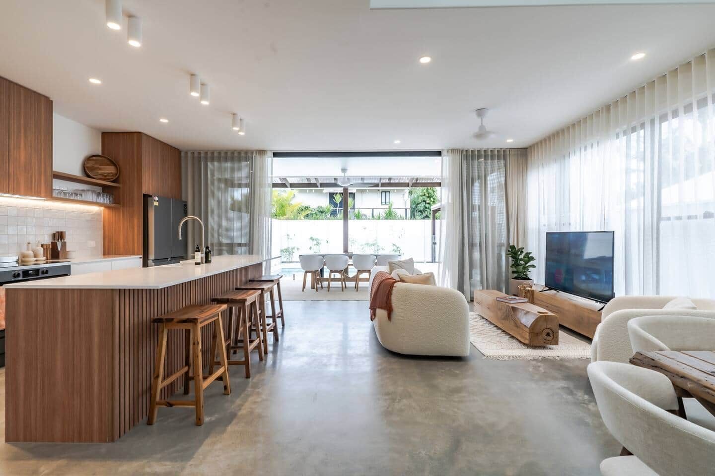 Una cocina moderna con una isla central, taburetes de bar, un comedor con mesa y sillas, un sofá y un televisor.