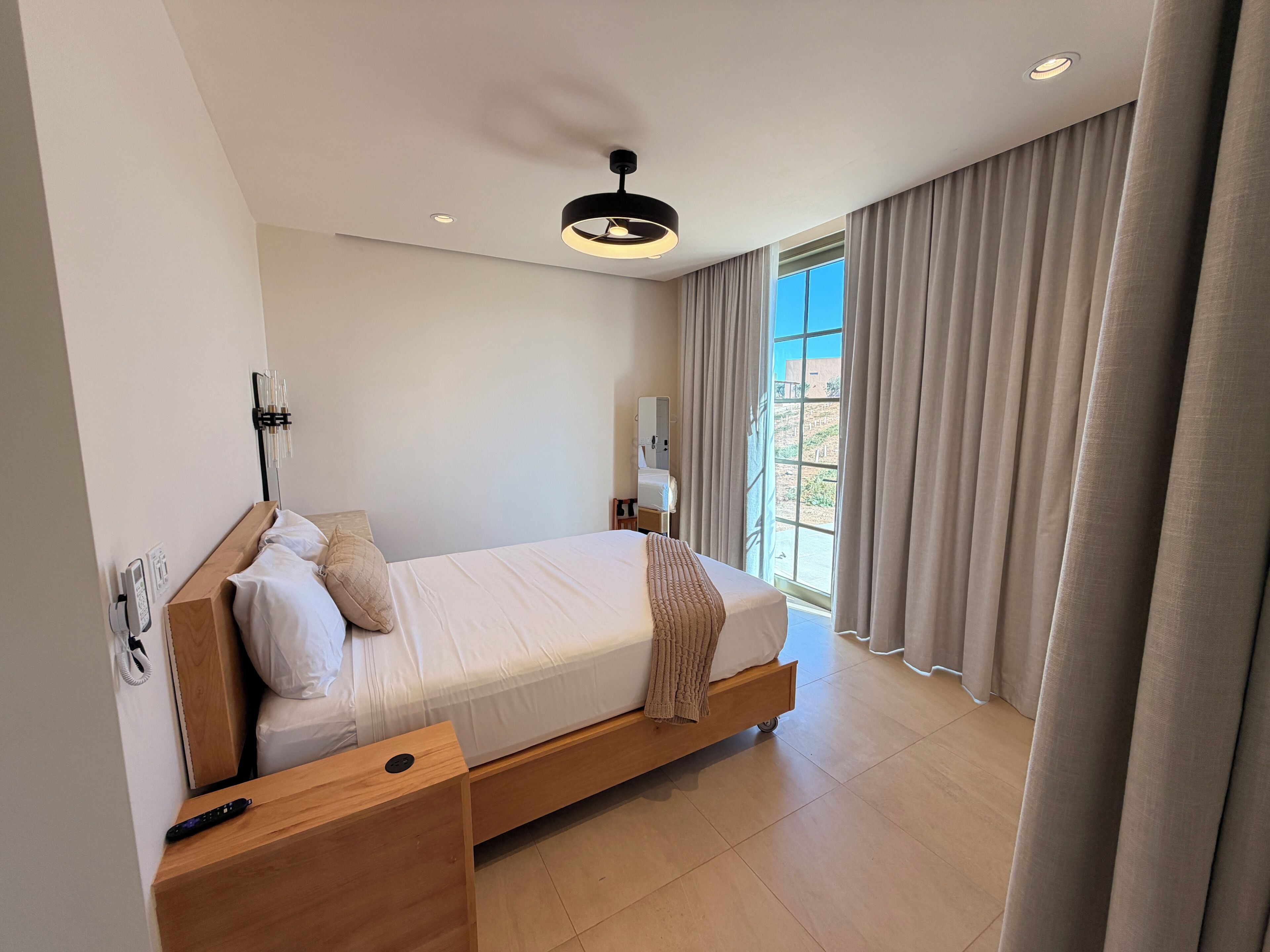 Una habitación de hotel moderna con una cama de madera, una mesita de noche, un control remoto y una ventana con cortinas.
