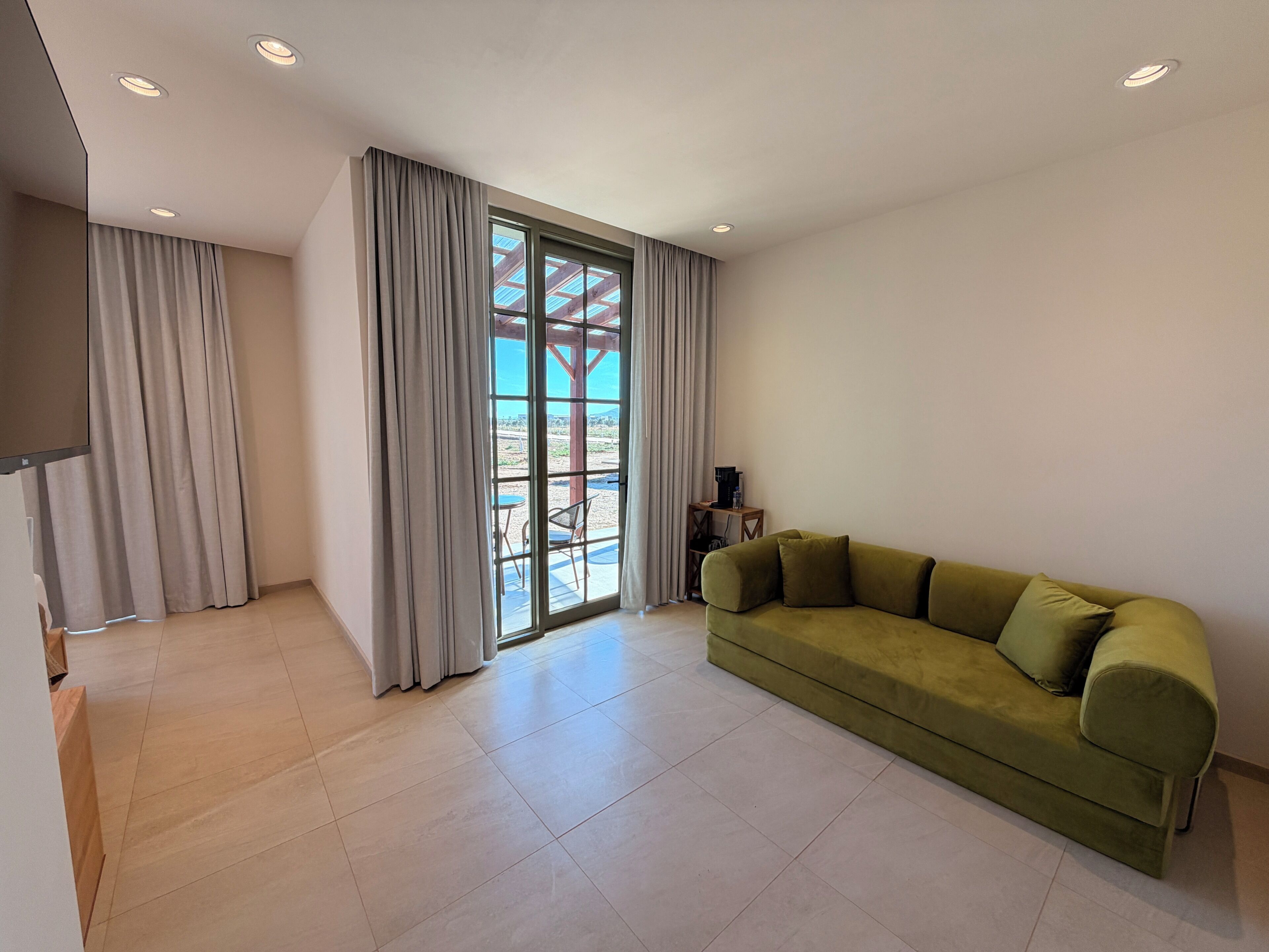 Un salon moderne avec un canapé vert, une grande porte-fenêtre coulissante donnant sur un espace extérieur et un éclairage encastré au plafond.