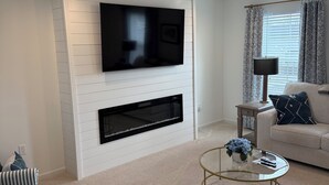 A living room with a flat-screen TV mounted on a white wall, a fireplace below it, a round coffee table, and a sofa with patterned cushions. - The Nittany House in Bellfonte (Bellefonte)
