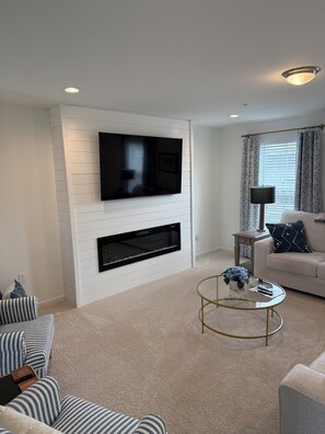 A living room with a flat-screen TV mounted on a white wall, a fireplace below it, a round coffee table, and a sofa with patterned cushions. - The Nittany House in Bellfonte (Bellefonte)