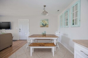 A dining area with a wooden table, white chairs, a bench, a painting, and a television on the wall. - 1501 - Carolina Breeze, Top-Floor 2BR Monteray Pines: Screened Balcony (Corolla)