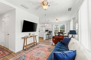 A spacious living room with a large flat-screen TV, a ceiling fan, and a dining area with a table and chairs. - Historic St Andrews Cottage Bikes, Walk to Marina, Porch, Patio & Yard (Panama City)