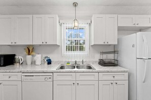 A kitchen with white cabinets, a double sink, a refrigerator, and a window with a view of trees. - Historic St Andrews Cottage Bikes, Walk to Marina, Porch, Patio & Yard (Panama City)