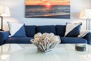 Ein modernes Wohnzimmer mit einer Couch, einem Glastisch, Dekorationselementen und einem Sonnenuntergangsbild an der Wand.