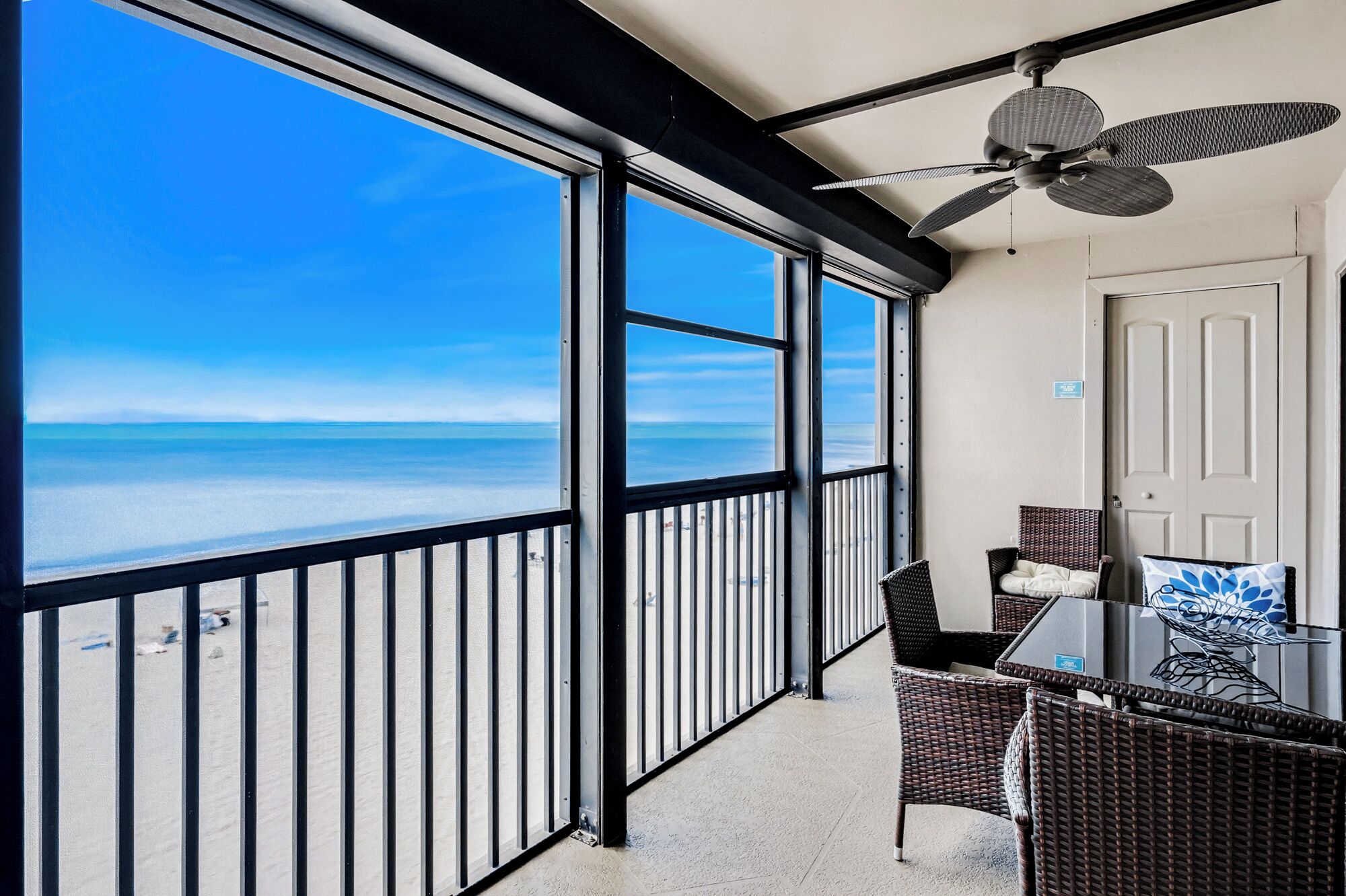 Varanda com vista para a praia, ventilador de teto, móveis de vime e uma porta.