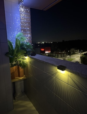 Ein Balkon mit einem Blumentopf, Lichterketten und Blick auf die beleuchtete Stadt bei Nacht.