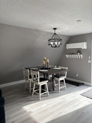 A dining area with a round table, white chairs, a chandelier, an air conditioner, and a sign on the wall.