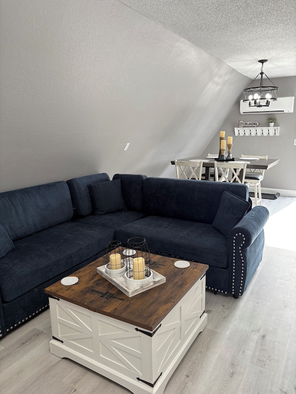 A modern living room with a dark blue sectional sofa, a wooden coffee table with candles, a dining area with a white table and chairs, and a ceiling light fixture.