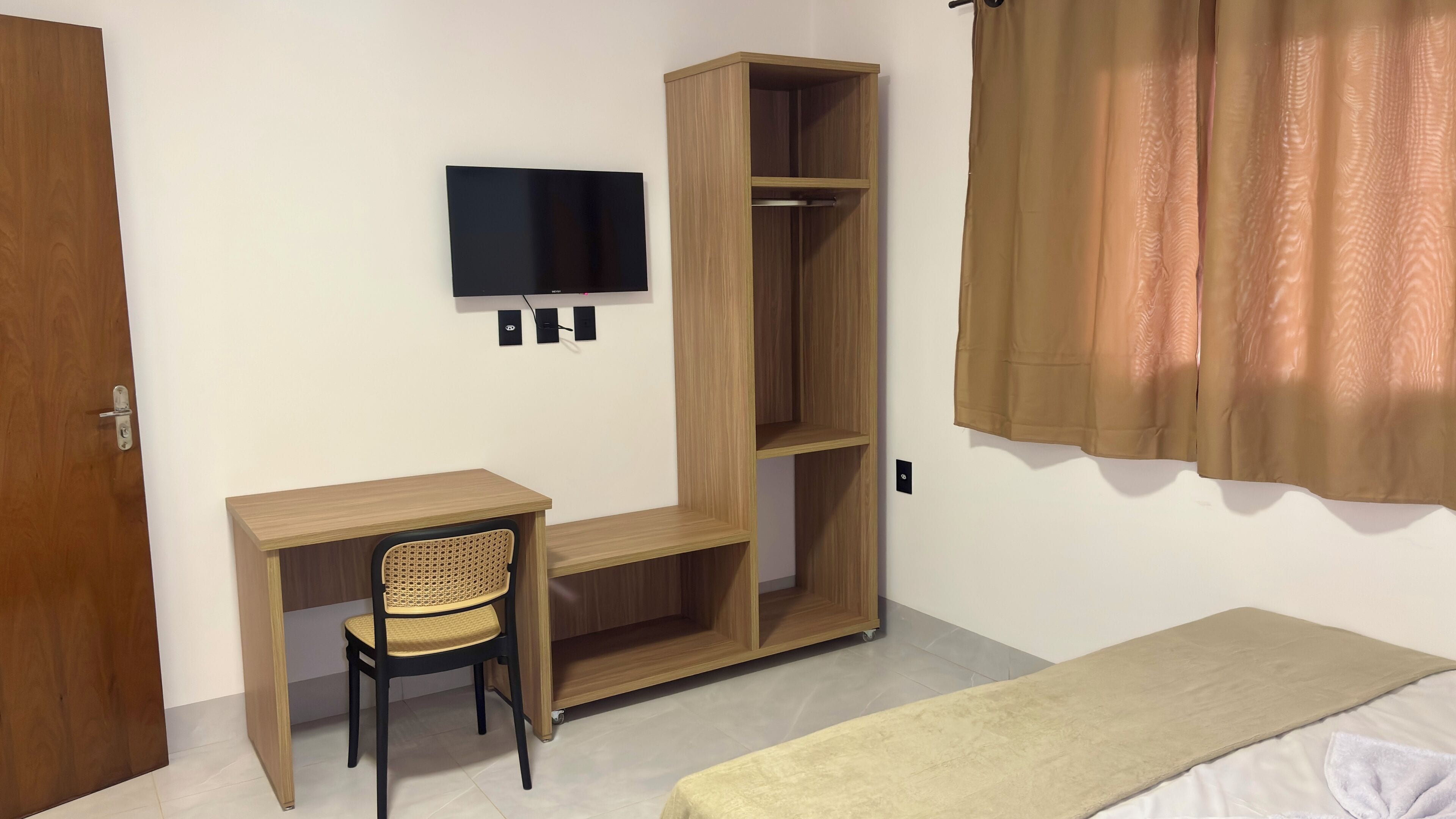 A room with a wooden desk, a chair, a TV mounted on the wall, a wardrobe, a bed, and a curtain.
