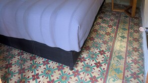 A bed with a grey and white duvet, a wooden side table with a lamp, and a patterned floor. - A hotel in a café (Oaxaca de Juárez)