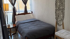 A bedroom with a bed, a wooden nightstand, a mirror, a window with curtains, a radiator, and a wall-mounted ironing board. - A hotel in a café (Oaxaca de Juárez)