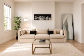 A living room with a beige sofa, a glass coffee table, a floor lamp, a cityscape painting, and a large mirror. - 15 min to NYC, Spacious & Private 3BR Home Sleeps 12 (Jersey City)