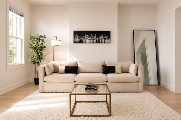 Un salon avec un canapé beige, une table basse en verre, un lampadaire, un tableau représentant un paysage urbain et un grand miroir.