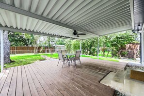 Eine überdachte Terrasse mit Tisch und Stühlen, einem Deckenventilator und Blick auf einen Garten mit Holzzaun und Bäumen.