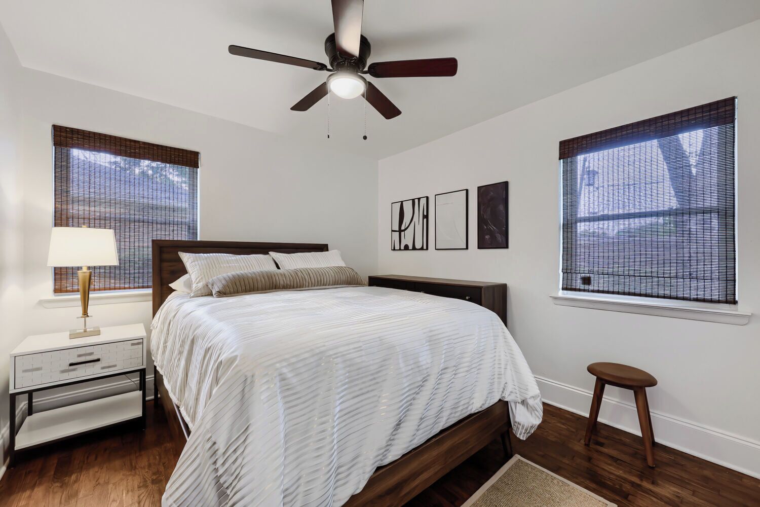 A bedroom with a bed, a nightstand, a lamp, a ceiling fan, and two windows with blinds.