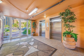 A hotel lobby with a tiled floor, a reception desk, a potted plant, and an elevator.