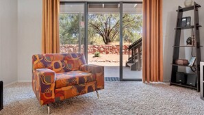 A room with a patterned armchair, a bookshelf, and a sliding glass door leading to an outdoor area with a view of trees and a stone wall.