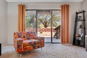 Ein Zimmer mit einem gemusterten Sessel, einem Bücherregal und einer Glasschiebetür zu einem Außenbereich mit Blick auf Bäume und eine Steinmauer.