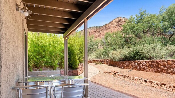 An outdoor patio with metal tables and chairs, a stone wall, and a view of a rocky hillside with greenery.