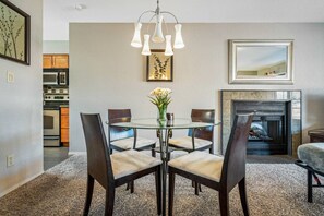 Een eethoek met een glazen tafel, vier stoelen, een vaas met bloemen, een open haard en een keuken op de achtergrond.