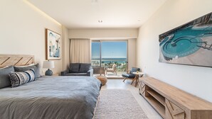 Una habitación de hotel moderna con una cama grande, un área de descanso con vista al océano, una mesa de comedor y un cuadro de gran tamaño.