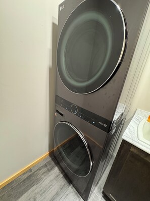 A black LG washing machine with a control panel and a glass door, placed in a room with a tiled floor and a white toilet. - Black Bear Ridge – Cozy 2BR + Loft Cabin 200 Private Acres Near Ellicottville NY (Franklinville)