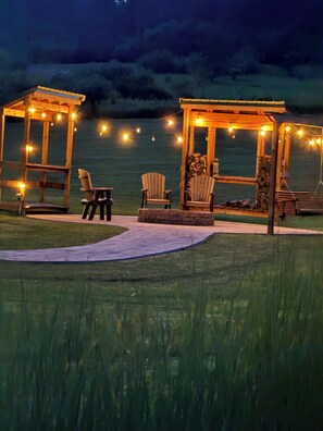 An outdoor seating area with wooden chairs and a pergola, surrounded by a grassy lawn and a body of water. - Black Bear Ridge – Cozy 2BR + Loft Cabin 200 Private Acres Near Ellicottville NY (Franklinville)