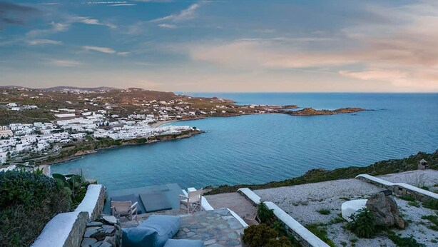 Un pueblo costero con edificios blancos, un camino de piedra, mesas al aire libre y vista al mar.