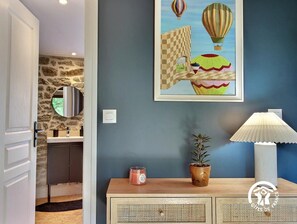 A room with a stone wall, a mirror, a painting of hot air balloons, a potted plant, a lamp, and a wooden cabinet. - La Grée du Moulin - PEAULE (PEAULE)