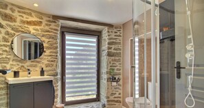 A bathroom with a stone wall, a round mirror, a sink with a faucet, a toilet, and a glass-enclosed shower. - La Grée du Moulin - PEAULE (PEAULE)