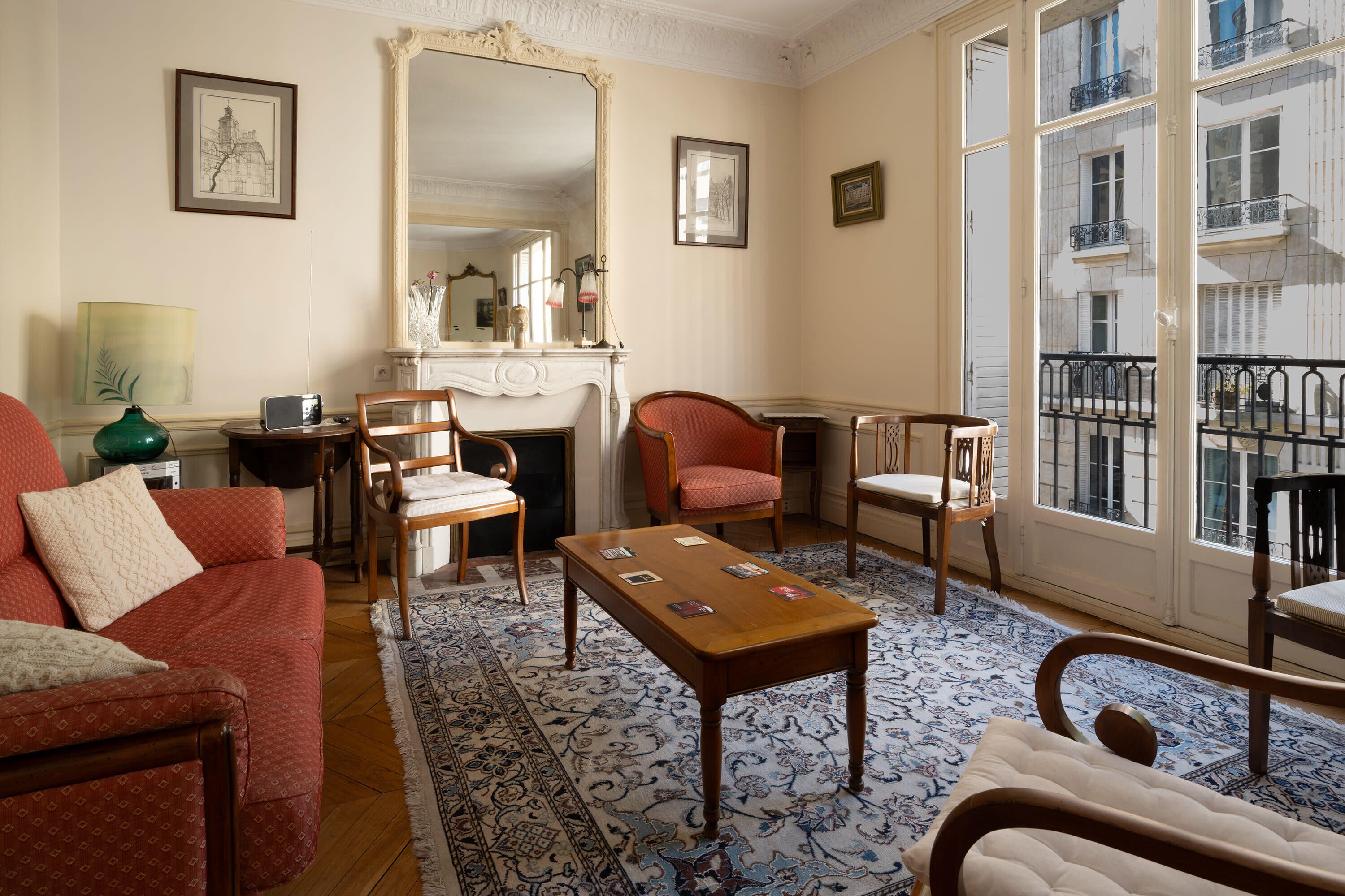 Un salon comprenant un canapé, des fauteuils, une table basse, une cheminée et de grandes fenêtres donnant sur des immeubles.