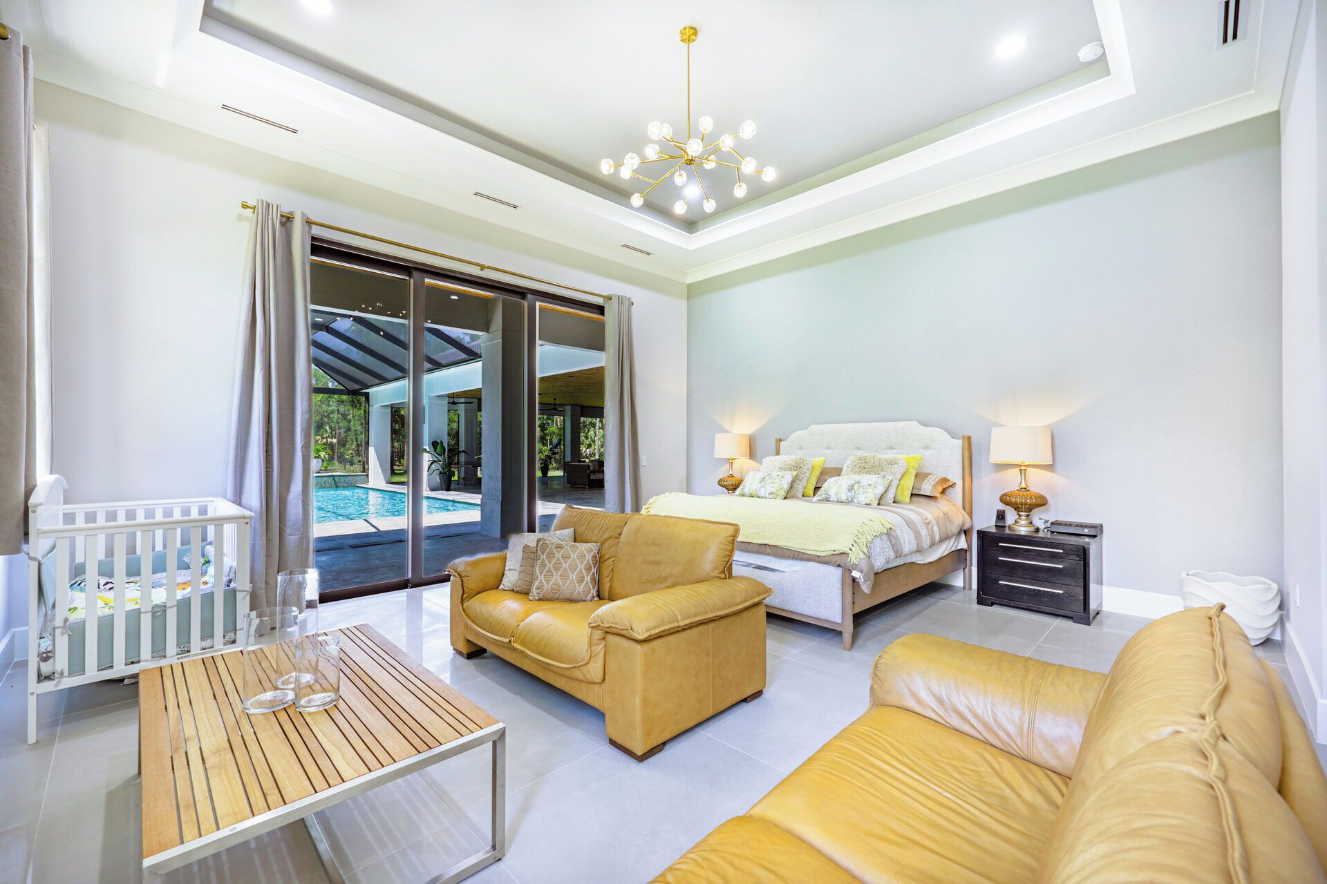 A modern bedroom with a bed, bedside tables, a sofa, a baby crib, and a view of a swimming pool.