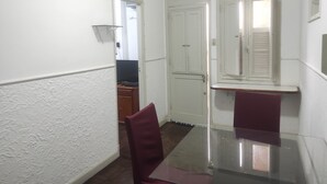 En smal hall med en vit dörr, ett litet fönster med fönsterluckor och ett glasbord med två stolar.