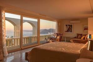 A hotel room with a large bed, a balcony with a view of a bridge and mountains, a small table with chairs, and a painting on the wall. - Santorini Gem, Santorini Gem (Thira - Santorini)
