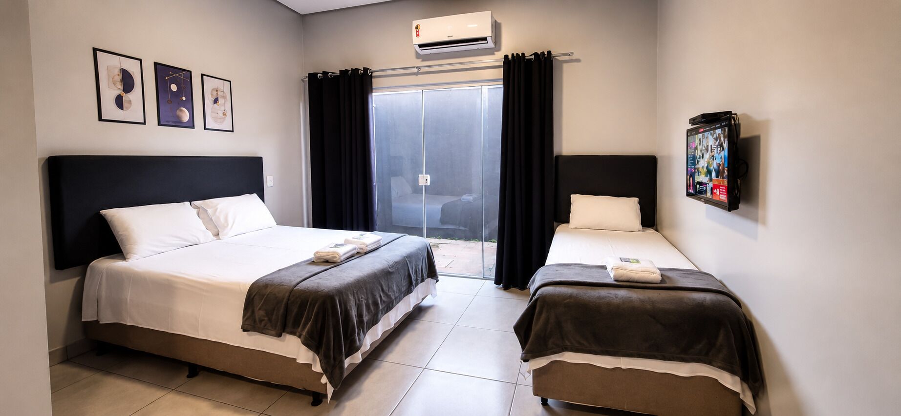 Een hotelkamer met twee bedden, een groot raam met gordijnen, een airconditioner en een bevestigde televisie.