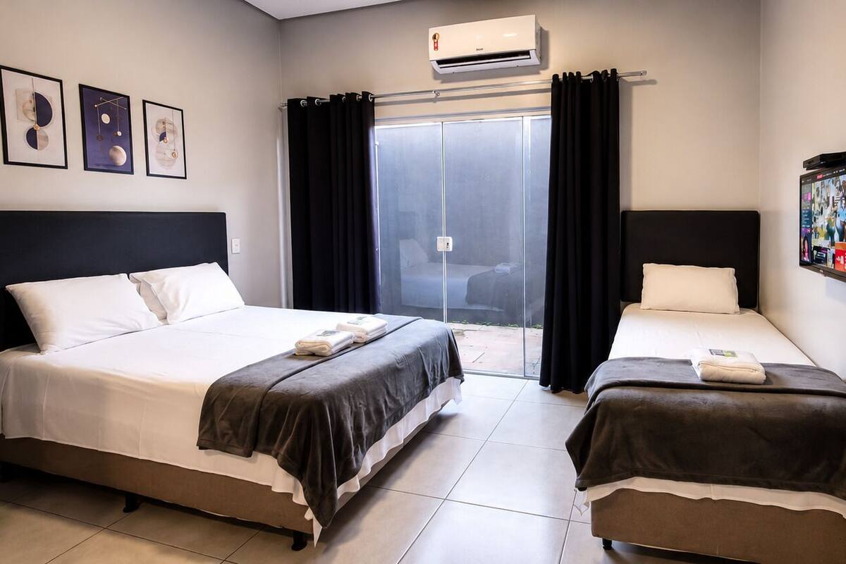 A hotel room with two beds, a large window with curtains, an air conditioning unit, and a mounted TV.