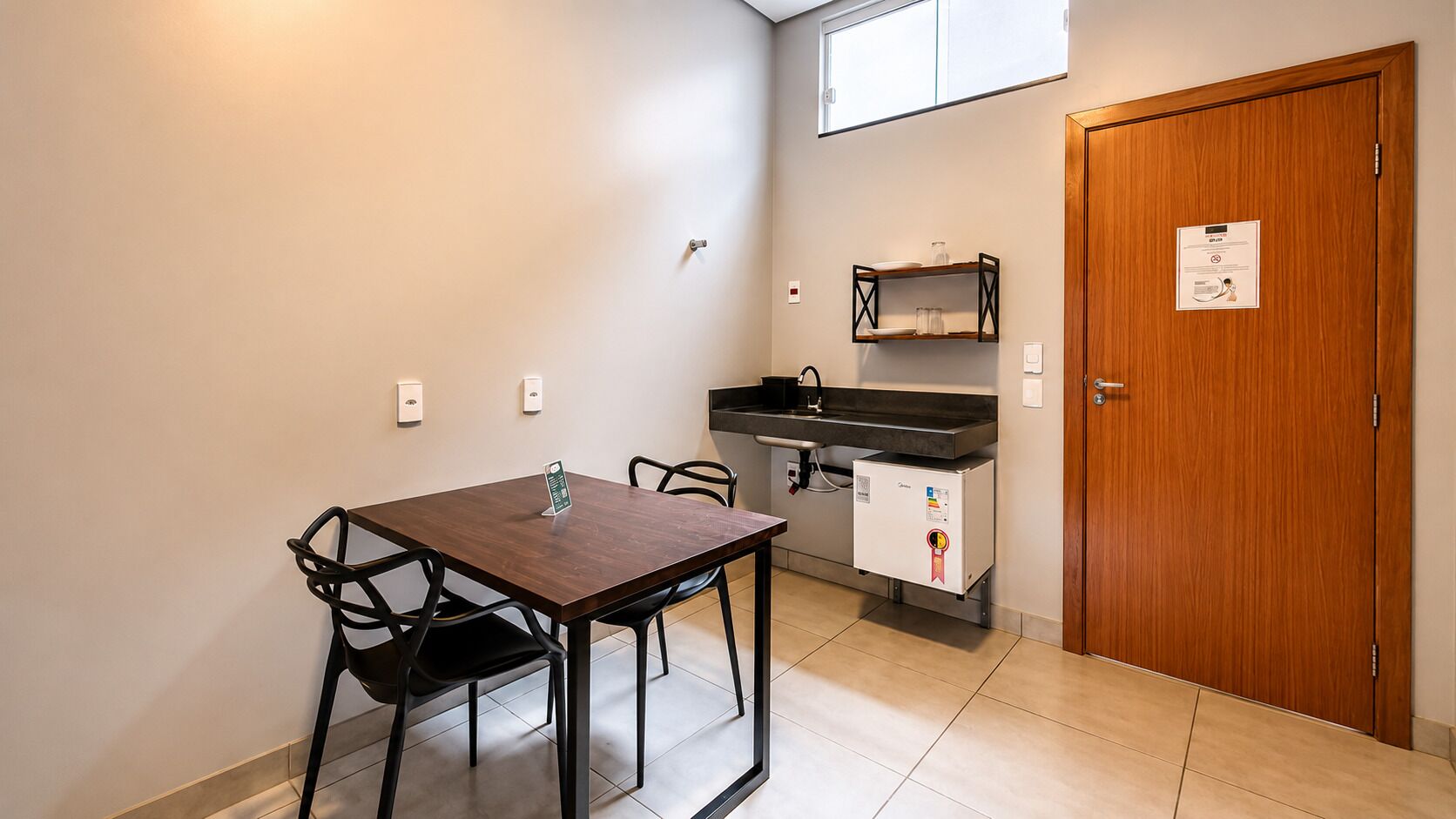 A small kitchenette with a refrigerator, a sink, and a microwave. There is a wooden door and a window above the sink.