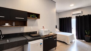 Uma cozinha compacta com micro-ondas, pia e armários, uma cama com cobertura branca e um aparelho de ar condicionado.