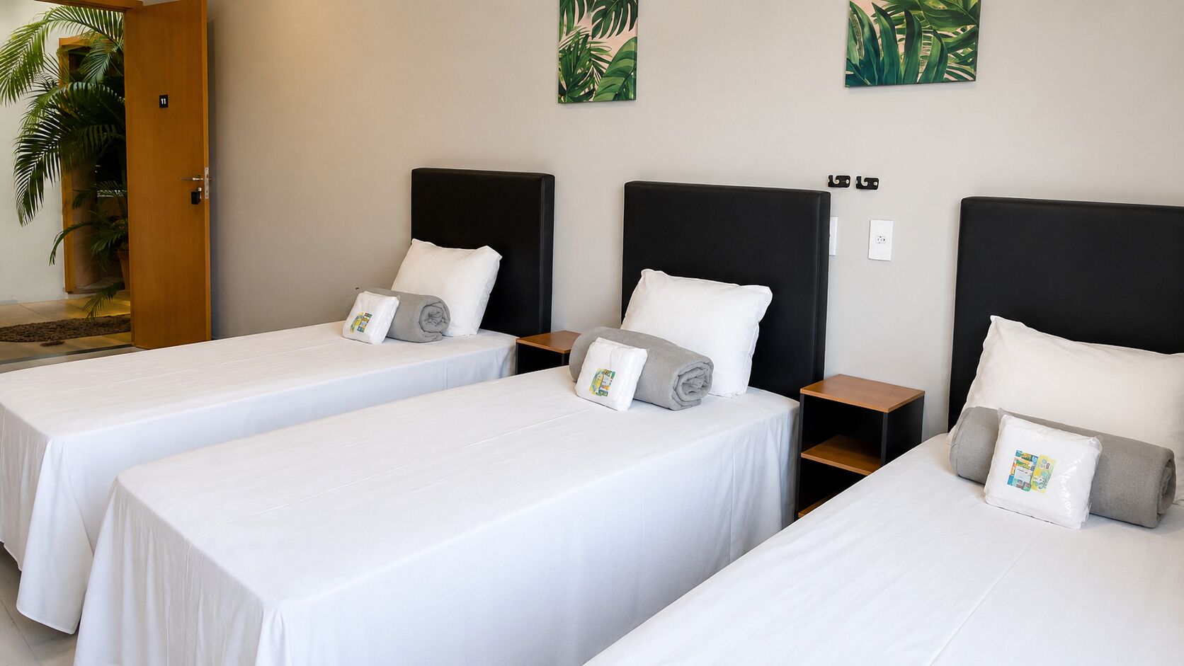 Een hotelkamer met drie enkele bedden, elk met een zwart hoofdbord, witte beddengoed en een klein houten nachtkastje.