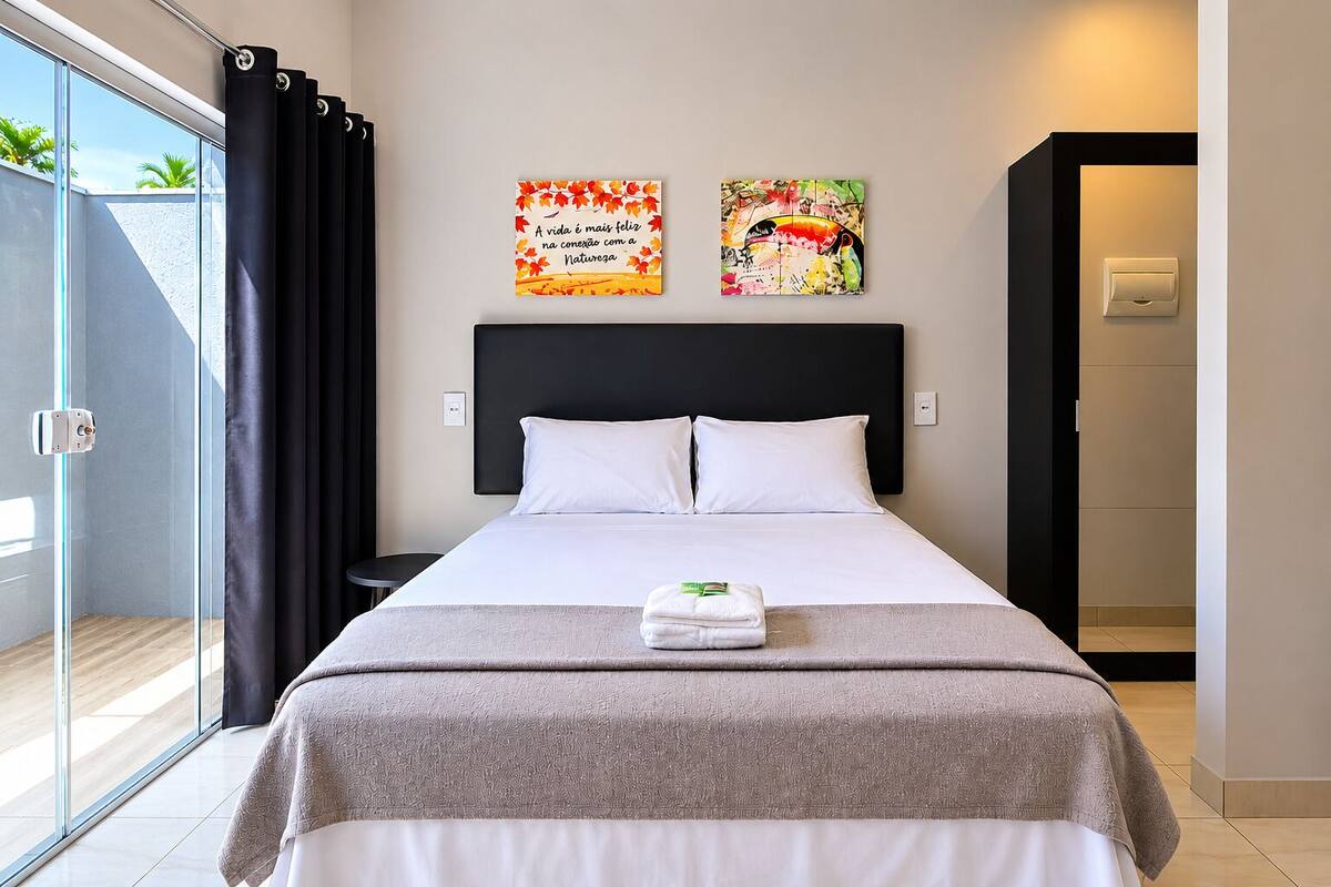 A hotel room with a bed, two wall art pieces, a window with curtains, and a door to the bathroom.